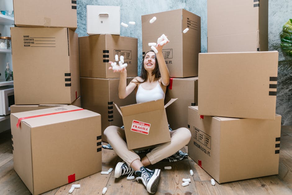 A young woman with long dark hair and a white sleeveless top is sitting on a wooden floor surrounded by multiple large cardboard moving boxes, some labeled with handling instructions. She is smiling and throwing white packing foam peanuts into the air, with a partially open box on her lap. The boxes vary in size and are stacked against a textured blue and gray wall, indicating an indoor setting likely prepared for a home relocation or furniture transport. To the left, a small portion of a kitchen counter with a bottle is visible, and in the background, a green plant adds a touch of decor. The scene captures the packing process involved in house removals, with equipment such as foam peanuts used for fragile item protection, reflecting the logistical aspects of professional removals services offered by Man With a Van Cowley, who specialise in house moving and packing support.