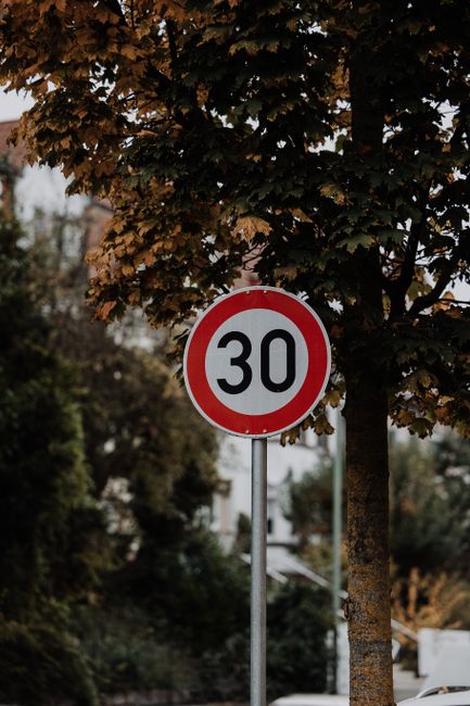 A round traffic sign displaying a speed limit of 30 miles per hour, mounted on a metal pole situated on a street surrounded by greenery. In the background, a mature tree with dense, brownish-green leaves partially obscures the sign, with additional trees and residential buildings visible further behind. The scene is captured during daylight hours, with natural lighting highlighting the sign and the surrounding environment. This setting relates to the context of house removals and local road regulations that require awareness of legal limits during moving or furniture transport, as indicated on the webpage about council fines and legal limits for Cowley house removals by Man With a Van Cowley.