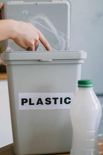 A person’s hand is seen placing a clear plastic container into a grey plastic recycling bin labeled 'PLASTIC' with black text in a home environment. The bin is situated on a flat surface indoors, with a partly visible white plastic bottle with a green cap standing in front of it. The background is blurred, emphasizing the action of sorting recyclable plastic materials during packing or decluttering in preparation for a house removal. The scene reflects responsible disposal and recycling practices associated with home relocation and furniture transport services provided by Man With a Van Cowley, supporting efficient move and waste management processes.