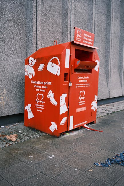 A red donation and recycling point mailbox positioned on a concrete pavement outside a building with a grey wall. The mailbox has a rectangular opening for donations, along with a labeled slot for clothes, books, and other items, surrounded by white graphic icons related to charity donations such as a teddy bear, handbag, shoes, and folded clothes. There is a small sign on top encouraging community support. The mailbox appears to be part of a house removal or moving logistics setup, with nearby debris including a blue plastic bag on the ground. The scene is well-lit with natural daylight, and the mailbox is associated with charity collection efforts linked to community recycling options for removals, as featured on the Man With a Van Cowley website.