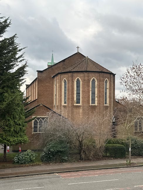 A large brick church building with tall, narrow stained glass windows arranged vertically along its curved exterior wall. The church has a pointed, shingled roof with a small cross at the apex, and a green spire is visible behind it. In front of the church, there are leafless trees and neatly trimmed bushes along the sidewalk, with some low flowering plants near the base of the building. The area appears to be part of a residential or community district, with a road in the foreground and overcast sky overhead. The image, associated with Man With a Van Cowley, relates to house removals and furniture transport, suggesting a context of home relocation services near the Church Cowley and Cowley Road flats.