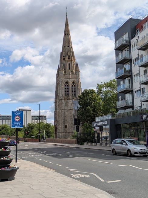 The image shows an urban street scene with a historic stone church featuring a tall, pointed spire and Gothic architectural elements in the background. To the right of the church, there are modern residential buildings with balconies and large windows. On the street, a silver car is parked along the curb, and a few other vehicles are visible further down the road. The sidewalk is lined with a small bike lane, marked by white lines and a bicycle symbol, and has decorative flower pots near the edge. A blue traffic sign indicating parking restrictions is also visible on the left side of the street. The sky is partly cloudy with patches of blue, and there are trees with green foliage along the sidewalk, adding a touch of nature to the scene. This setting provides a representative view of a typical cityscape where house removals or furniture transport, coordinated by companies like Man With a Van Cowley, could take place during home relocation or moving services.