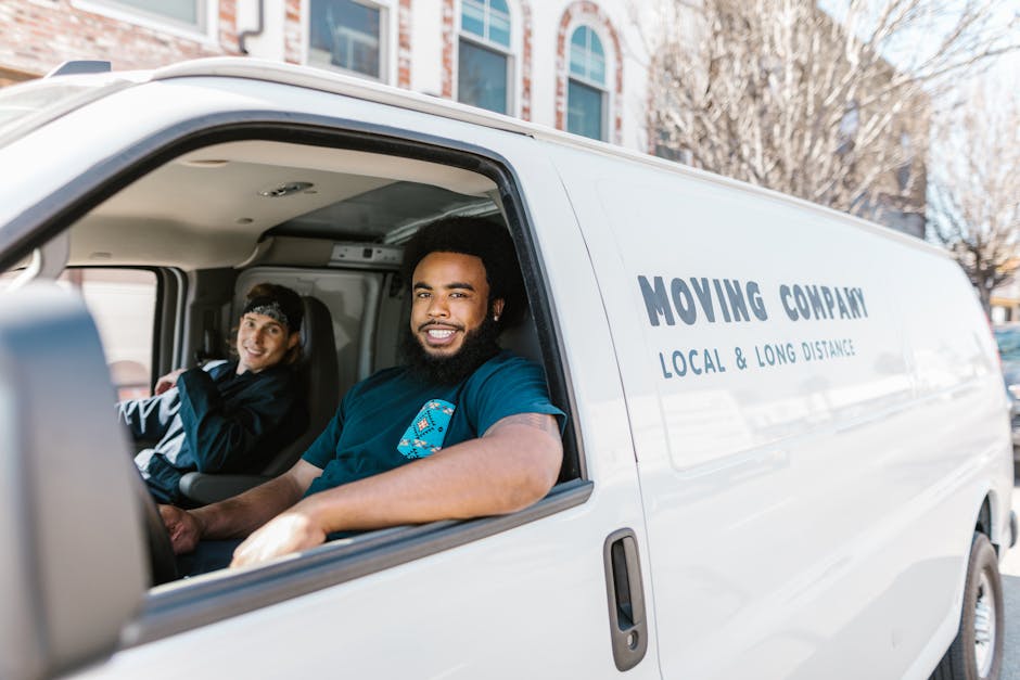 Inside the cab of a white moving van from Man With a Van Cowley, with two professional movers preparing for a house relocation. The driver, a man with a beard and short dark hair, sits in the driver's seat, smiling at the camera, wearing a navy blue T-shirt with a small graphic logo. Beside him, a woman with long dark hair, wearing a dark uniform and a headband, sits in the passenger seat, also smiling. The interior of the van features a clean dashboard, and outside the open driver's side window, residential buildings and trees are visible during daytime. The van is marked with black text on the side that reads 'MOVING COMPANY, LOCAL & LONG DISTANCE,' indicating a professional furniture transport service. This scene captures the loading and transport process involved in home relocation, with the focus on preparing for a move that includes packing, lifting, and loading furniture and boxes into the vehicle for either local or long-distance removals.
