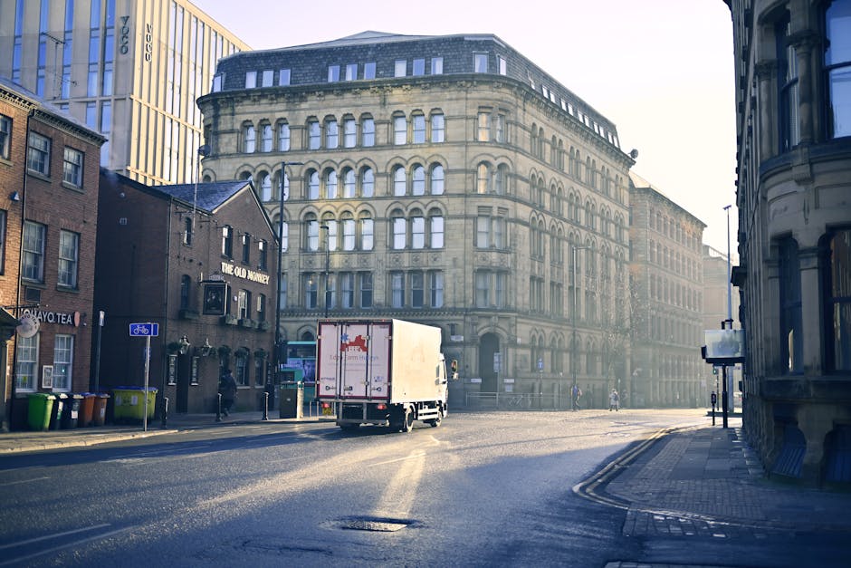 A white moving truck with red markings is parked on a wide, paved city street in Cowley, with the rear doors open, positioned near the entrance of a multi-story historic building with arched windows and ornate stone detailing. The truck is surrounded by several cardboard boxes, plastic-wrapped furniture, and packing materials arranged on the pavement, indicating an ongoing home relocation process. Nearby, a trolley and moving blankets are visible, likely used for furniture transport and protection during the loading process. To the left, a small pub named 'The Old Monty' is situated in a dark brick building, with a row of green and orange wheelie bins lined up outside. Pedestrians and cyclists are seen in the background, with some walking along the sidewalk. The lighting suggests early morning or late afternoon, casting long shadows and creating a slightly hazy atmosphere, characteristic of city environments involved in packing and moving activities. Man With a Van Cowley is involved in the furniture transport and house removals occurring in this urban setting.