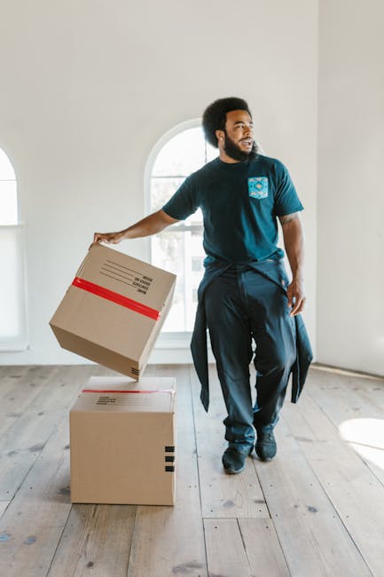 Inside the cab of a white moving van from Man With a Van Cowley, with two professional movers preparing for a house relocation. The driver, a man with a beard and short dark hair, sits in the driver's seat, smiling at the camera, wearing a navy blue T-shirt with a small graphic logo. Beside him, a woman with long dark hair, wearing a dark uniform and a headband, sits in the passenger seat, also smiling. The interior of the van features a clean dashboard, and outside the open driver's side window, residential buildings and trees are visible during daytime. The van is marked with black text on the side that reads 'MOVING COMPANY, LOCAL & LONG DISTANCE,' indicating a professional furniture transport service. This scene captures the loading and transport process involved in home relocation, with the focus on preparing for a move that includes packing, lifting, and loading furniture and boxes into the vehicle for either local or long-distance removals.