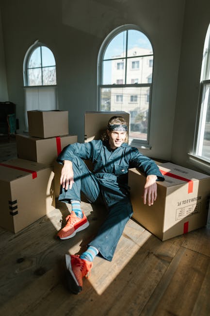 A young man sitting on the wooden floor inside a sunlit room with large arched windows, surrounded by several closed cardboard boxes sealed with red packing tape, suggests he is in the process of packing or preparing for a home relocation. He is dressed in casual working clothes, including orange athletic shoes with white soles, blue jeans, and a dark jacket, and is wearing a grey cap with a pattern. The room's natural light highlights the boxes and the man's relaxed posture, indicating a moment during furniture transport, packing, or moving logistics. The windows offer a view of nearby buildings, and the clean, minimally furnished space reflects an active move or packing process supported by professional removals services such as those provided by Man With a Van Cowley.