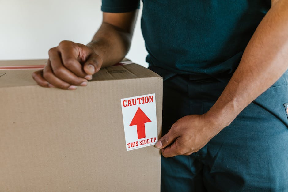 A person wearing a dark green T-shirt and dark trousers is holding a medium-sized cardboard box during a home relocation process. The box has a white and red caution label with an upward-pointing arrow and the text 'CAUTION THIS SIDE UP'. The individual’s hands are gripping the box firmly, one hand near the top and the other supporting from below. The background shows an indoor environment with a plain white wall, indicating they are inside a property, preparing for furniture transport and packing during a same day removal. This loading process involves careful handling of packed items, with the box likely containing household goods or fragile items. The scene captures the logistics of loading and moving boxes, possibly onto a van, as part of the furniture transport and packing and moving services provided by Man With a Van Cowley, ensuring efficient house removals and minimal delays in relocation.