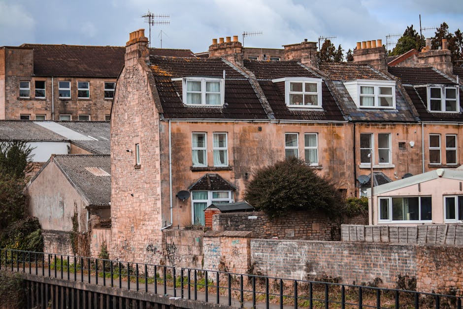 The image shows a row of old terraced houses constructed from reddish-brown brick, situated in a residential area with a small fence at the forefront. The houses feature pitched roofs covered with dark tiles, with several roof windows and chimneys visible on the rooftops. The windows are mostly white-framed, double-glazed, with some featuring curtains. In front of the houses, a brick wall partially surrounds the property, with a large bush growing near the wall, obscuring part of the lower windows. A satellite dish is mounted on one house’s wall. Visible elements indicate an outdoor area that may be used during home removals or furniture transport, with adjacent buildings appearing to be part of the same residential block. The sky above is partly cloudy, and the lighting suggests daytime, suitable for a home relocation process. The context implies a typical residential street scene, aligning with house removals and moving services provided by Man With a Van Cowley.