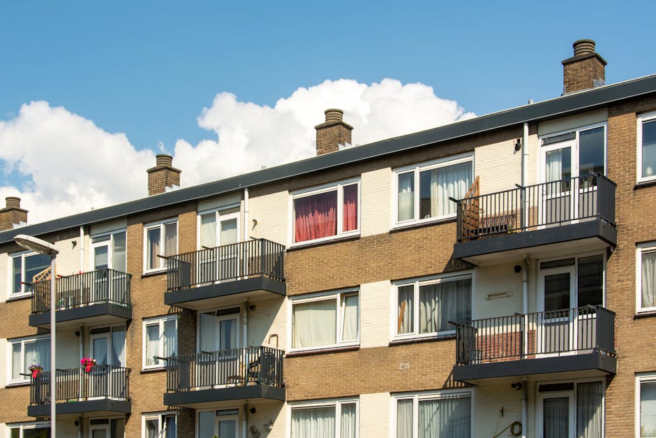 Photograph of the exterior of a three-story residential building with multiple balconies, situated in a residential area. The building features a brick facade with horizontal white sections, each with several large windows, some with curtains or blinds visible. Four of the balconies, equipped with black metal railings, are visible; the balconies contain various items such as potted plants, outdoor furniture, or blankets. On the top floor, a balcony has a wooden chair, and some windows have curtains in different colours like red and white. The building is illuminated by natural daylight, with a bright blue skyic background, dotted with white clouds. The setting suggests daytime, suitable for house removals or furniture transport activities, aligning with professional moving services like those offered by Man With a Van Cowley, who specialise in home relocation and packing and moving processes.