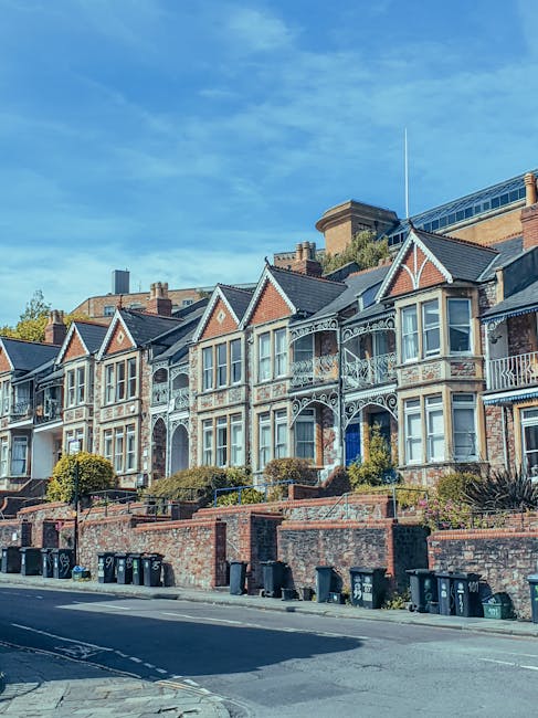 The image shows a row of old terraced houses constructed from reddish-brown brick, situated in a residential area with a small fence at the forefront. The houses feature pitched roofs covered with dark tiles, with several roof windows and chimneys visible on the rooftops. The windows are mostly white-framed, double-glazed, with some featuring curtains. In front of the houses, a brick wall partially surrounds the property, with a large bush growing near the wall, obscuring part of the lower windows. A satellite dish is mounted on one house’s wall. Visible elements indicate an outdoor area that may be used during home removals or furniture transport, with adjacent buildings appearing to be part of the same residential block. The sky above is partly cloudy, and the lighting suggests daytime, suitable for a home relocation process. The context implies a typical residential street scene, aligning with house removals and moving services provided by Man With a Van Cowley.