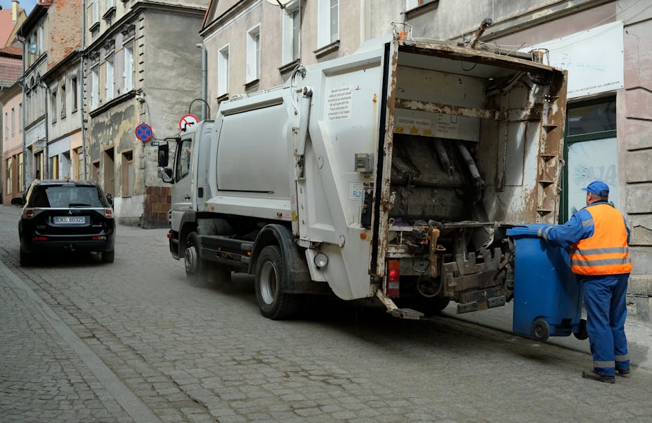 A waste collection truck operated by Man With a Van Cowley is positioned on a cobblestone street in an urban residential area, with its rear compartment open, revealing metal mechanical components. The truck is white and shows signs of rust and dirt at the rear. A worker wearing a blue uniform, high-visibility orange vest, and a blue cap is seen placing a blue wheeled bin into the truck's open compartment, part of a home relocation or busy furniture transport process involving waste removal. On the street, there is a black car parked nearby, and surrounding buildings with multi-story facades, some displaying worn plaster and brickwork, are visible along with sidewalk signs indicating parking restrictions. The outdoor setting is daytime with natural light, and the scene captures a typical rubbish collection activity in an urban environment, relevant to residential waste management and removal services performed by Man With a Van Cowley.