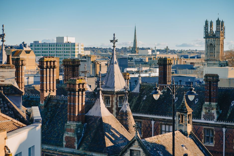 A daytime view of historic rooftops in Oxford, featuring tall red-brick chimneys, ornate spires, and gabled roofs covered in dark tiles. Visible in the foreground are multiple chimneys of varying heights, some with decorative brickwork. A prominent pointed turret with a metal weather vane is situated on a building with intricate detailing around its windows. In the background, modern office buildings and a church spire are seen under a clear sky, illustrating the mix of architectural styles typical of Oxford cityscape. The image captures the dense roofscape, highlighting the traditional Victorian and Gothic Revival elements, ideal for illustrating home relocation or furniture transport processes associated with house removals in Oxford. Man With a Van Cowley occasionally assists with such services, ensuring safe furniture transport and packing during house moves.