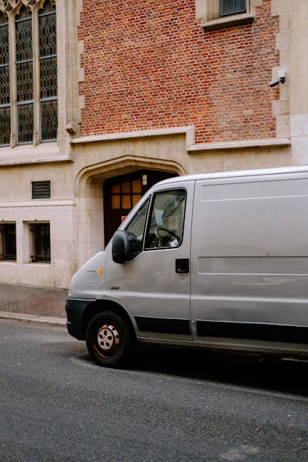 A grey panel van parked on a street in front of a historic brick building with large stained glass windows and a stone archway entrance. The van is positioned close to the curb, with the driver’s side visible, showing a side mirror and black wheel. Inside the vehicle, visible through the window, are cardboard boxes and packing materials, indicating a house removal or furniture transport process. Nearby, on the pavement, a hand truck or trolley may be partially visible, suggesting the ongoing loading or unloading of household items by staff from Man With a Van Cowley. The scene is well-lit with natural daylight, capturing an urban environment prepared for a residential move, with the van used for home relocation logistics and packing and moving services.