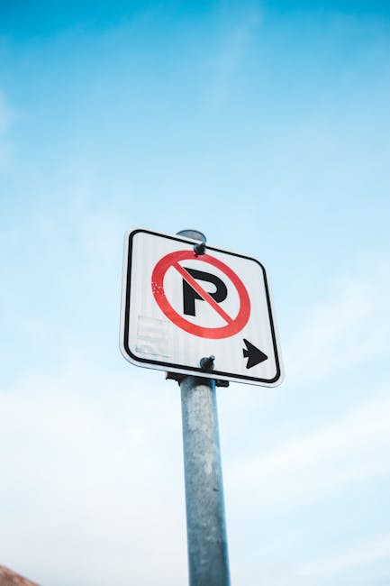 A vertical street sign mounted on a metal pole displays a white background with a red circle and diagonal line crossing out a black letter 'P', indicating no parking. Below the sign, there is a small black arrow pointing to the right. The background features a clear blue sky with some wispy white clouds. The sign is positioned at a height above ground level, suggesting it is placed to control parking restrictions in an area where house removals and furniture transport may occur. The image captures the sign clearly without any other objects or vehicles visible, providing a neutral and factual visual relevant to parking regulations for moving services such as those offered by Man With a Van Cowley.