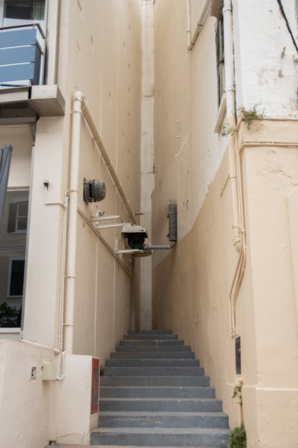 A narrow outdoor staircase made of grey concrete steps ascending between two tall, beige building walls. The left wall has visible white PVC pipes and a small metal conduit, with an electrical meter attached. The right wall features some weathering and a small sparse plant growing near the base. The area appears to be a confined service or alleyway, with limited space for manoeuvring. This setting likely depicts a location where Man With a Van Cowley facilitates home relocation or furniture transport through tight access points, such as narrow lanes or courtyards, highlighting challenges faced during packing, loading, and delivering household items in confined spaces during a house removal or moving service.