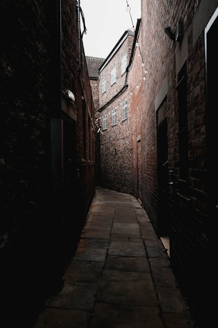 A narrow paved alleyway between two brick buildings, with the left side featuring a white-painted exterior wall and small, high-set windows, and the right side displaying a red-brick wall with a street sign reading 'FALLOWS LANE' attached near the top. The alley has a series of steps at the far end, leading up to a street with a lamppost, trees, and a partly cloudy blue sky overhead. This confined passage reflects a typical setting for house removals and furniture transport in tight access areas, as handled by Man With a Van Cowley, which specializes in residential moving services and packing logistics for home relocation projects.