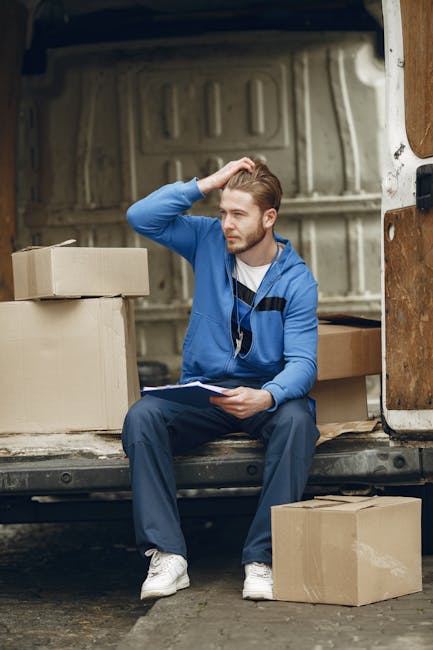 A young man with short, light brown hair and a beard is seated on the edge of an open moving van with a beige interior, surrounded by neatly packed cardboard boxes of various sizes. He is wearing a blue zip-up jacket, navy trousers, and white sneakers, and is holding a clipboard with a pen while scratching his head in a thoughtful manner. The van is parked on a paved surface, and the scene suggests a home relocation process involving packing, logistics planning, and preparing for furniture transport. The background shows a glimpse of the vehicle's internal side panel, indicating the loading area, with additional boxes inside the van. This image exemplifies the packing and loading stages of a professional removals service, with [COMPANY_NAME] providing the logistics for a house move in Cowley, as referenced in the page about removals booking delays.